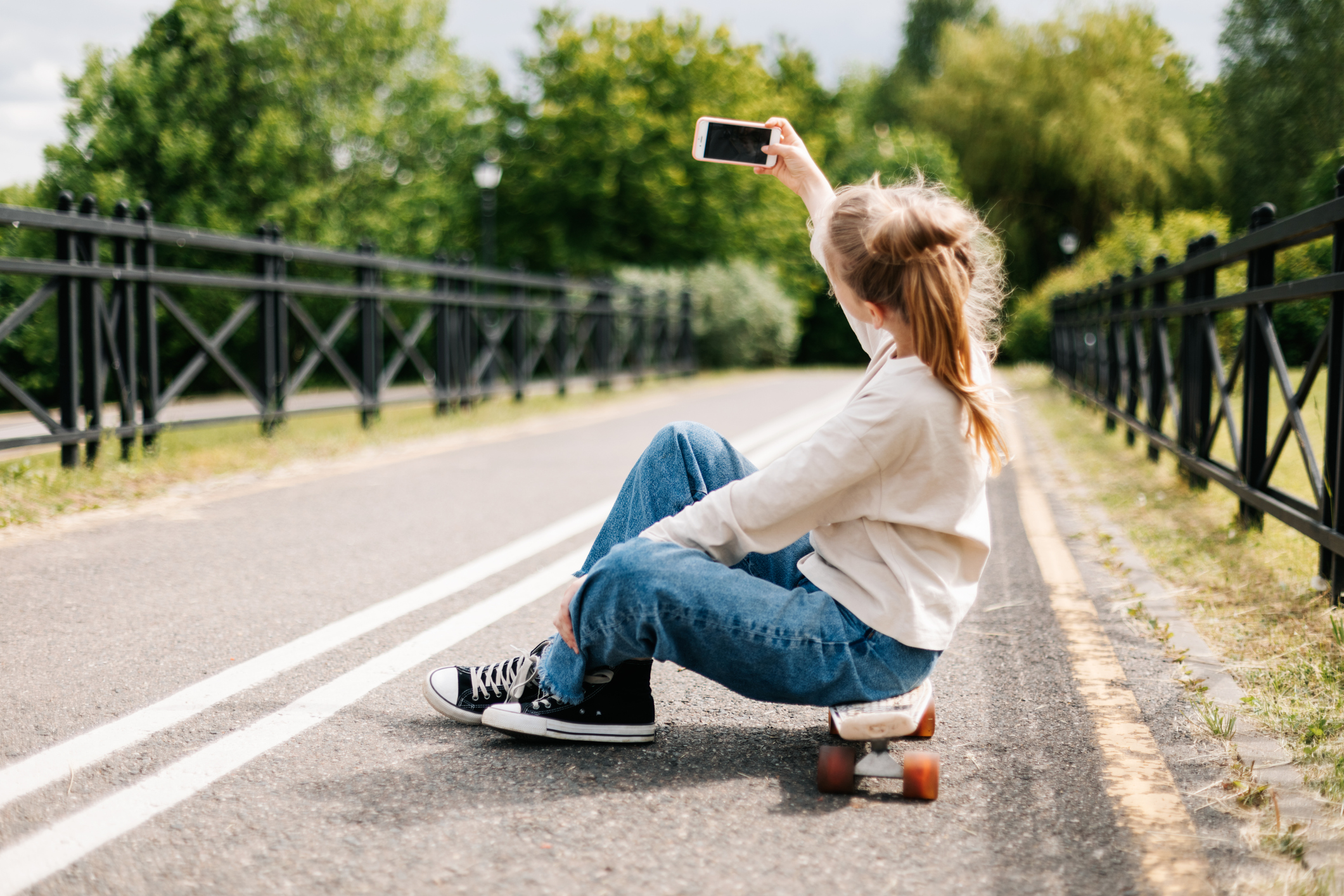 a young person sits on a skateboard on a road taking pictures with a smartphone