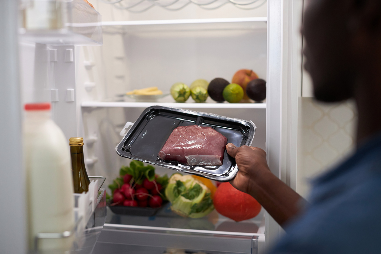 a person removing packaged meat from a fridge containing milk and vegetables