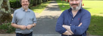 In this image there are two men standing outside in a park with trees and green grass behind them.