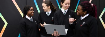 An image of four students in school uniforms, smiling and talking to each other. One is holding a laptop in their hand 