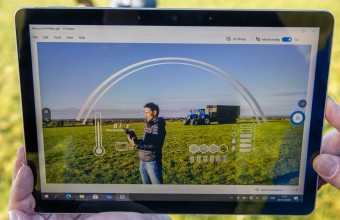 Hands hold a smart tablet with a green field in the background. On screen is an illustration of different information as a man stands in the field with farm vehicle in background.