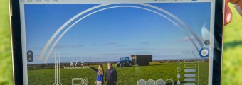 Hands hold a smart tablet with a green field in the background. On screen is an illustration of different information as two people stand in the field with farm vehicle in background.