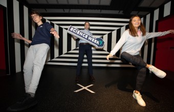 Three younger people standing in an illusions room holding a science week sign.