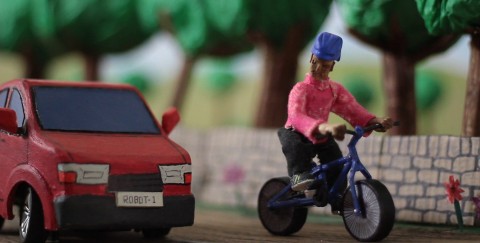 Clay model of a person cycling with a red car coming up beside them