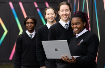 Image of 4 transition year students, wearing school uniforms, standing in front of a wall painted black with colourful stripes. One of the students is holding a laptop. 