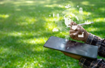 Person holding a smart tablet and pen; with green grass in the background and icons coming from the tablet representing various aspects of society