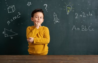 A child standing infront of a chalkboard with his arm crossed and hand on his face with a thinking expression on