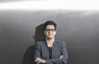 Image of a women with short dark hair with her arms folded with a grey background behind her.