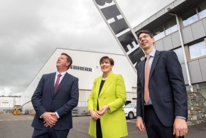 An image of two men in suit and one woman in a bright yellow coat standing in front of a building and looking up