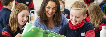 Image of a teacher reading a book with two pupils with many other pupils in the background.