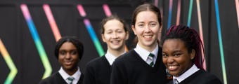 Image of 4 transition year students, wearing school uniforms, standing in front of a wall painted black with colourful stripes. One of the students is holding a laptop. 