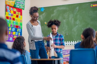 Imagine og young student holding a rocket at top of classroom with teacher.