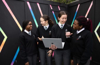 An image of four students in school uniforms, smiling and talking to each other. One is holding a laptop in their hand 