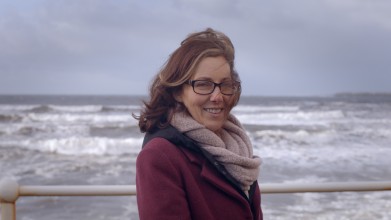 Rachel O'Mahony, a cancer survivors, is standing in front of the sea with waves in that background dressed in warm clothing.