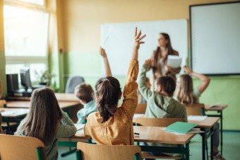 Picture of a teacher in a classroom with children with the arms raised to ask questions