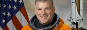 An image of a Colonel Greg "Box" Johnson in an orange NASA astronaut and holding his helmet in front of a USA flag and a space shuttle in the background