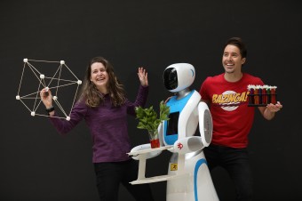 A picture of a robot holding a plant in between a woman in a purple top  and a man  in a red shirt