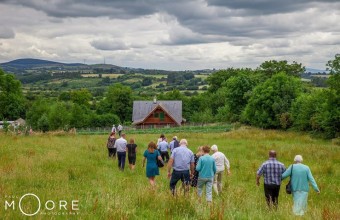 A group of people walking through a field towards a wooden farm house surrounded by fields and trees