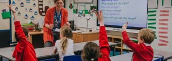 Young students with their hands up wearing a red uniform and teacher teaching them in the classroom.