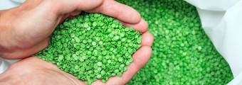 Hands holding green biodegradable plastic pellets above a bag of the same pellets. 