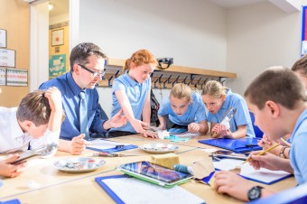 Teacher and students sit around tables and study a specimen in the middle of the table