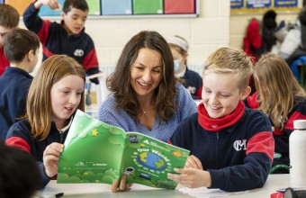 Image of a teacher reading a book with two pupils with many other pupils in the background.