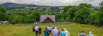 A group of people walking through a field towards a wooden farm house surrounded by fields and trees