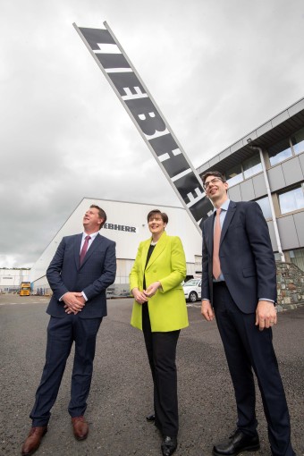 An image of two men in suit and one woman in a bright yellow coat standing in front of a building and looking up