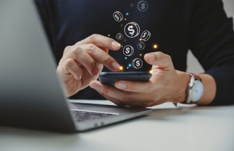 Person using smart phone. There are coin icons coming from the smart phone. There is also a laptop computer on the table in front of the person.