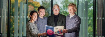 Picture of 3 males and 1 female standing in front of a glass background holding a book open.