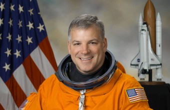 An image of a Colonel Greg "Box" Johnson in an orange NASA astronaut and holding his helmet in front of a USA flag and a space shuttle in the background