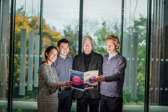 Four people standing beside each other holding a book in front of a glass door.