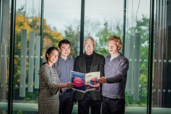 Four people standing beside each other holding a book in front of a glass door.